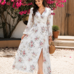 Woman wearing a white floral maxi dress in a sunlit garden, holding a woven handbag with pink flowers in the background.