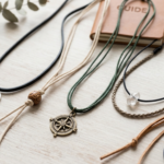 A variety of minimalist cord necklaces with different pendants, including a silver disc, a wooden bead, a bronze compass, and a raw crystal charm, arranged flat on a light wooden background next to a leather notebook and eucalyptus leaves.