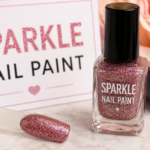 A close-up cosmetic scene featuring a glittery sparkle nail paint bottle with rose-gold shimmer polish and a matching swatch stick on a soft floral background.